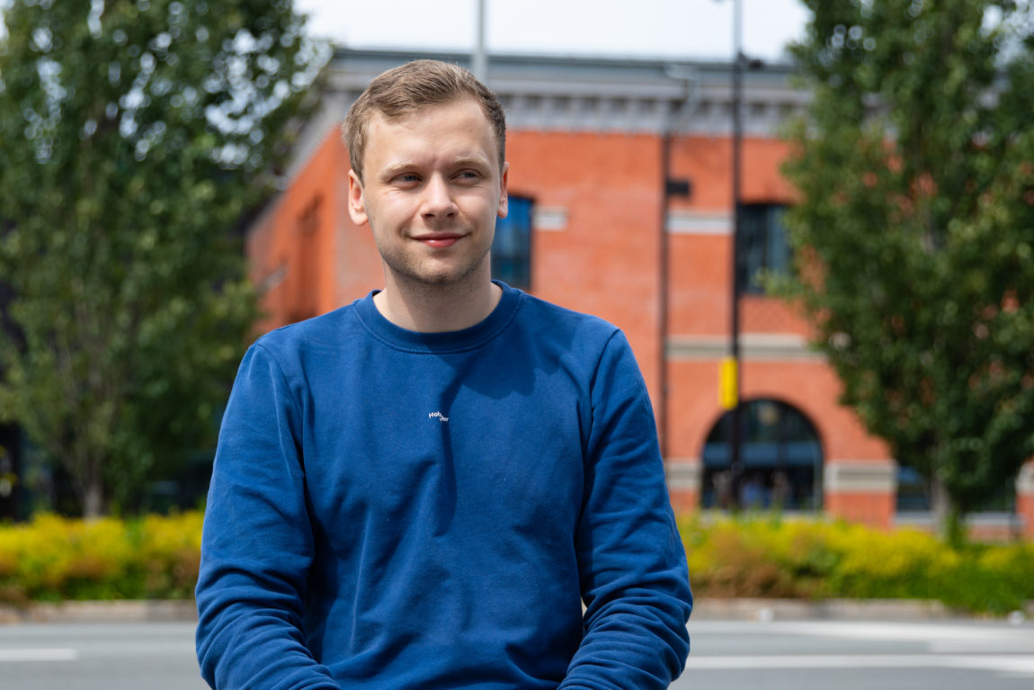 Sander Østlyngen sitter på en benk, og ser ut av bildet til høyre. Bak han er grønne trær og et oransje mursteinbygg. 