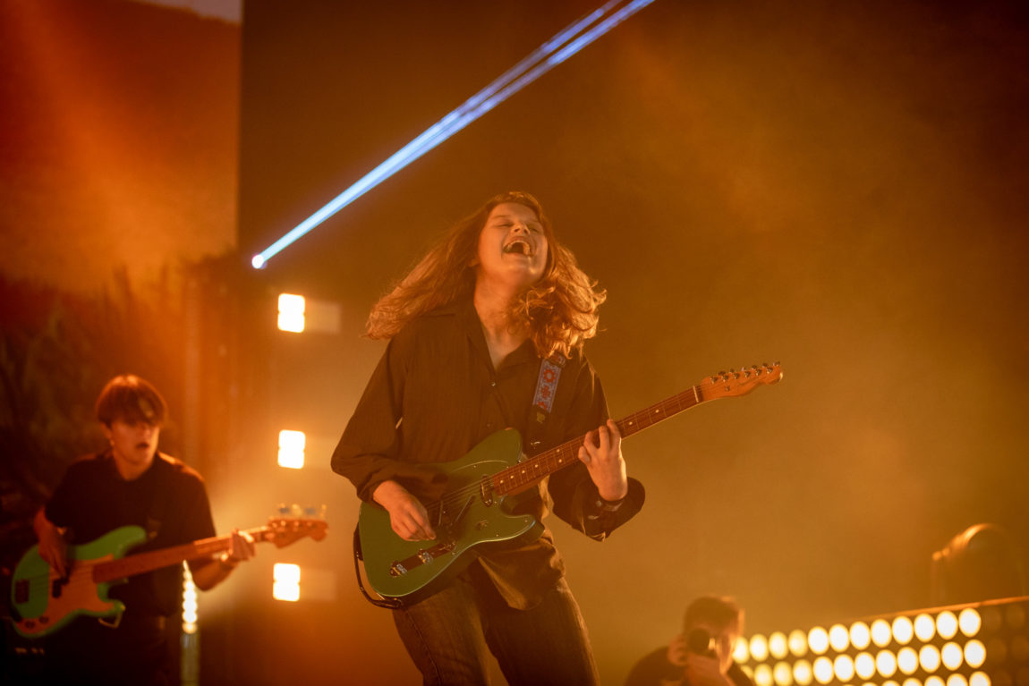 Girl in red spiller en rød gitar. Hun smiler mens håret henger ned. I bakgrunnen ser vi bassisten hennes.