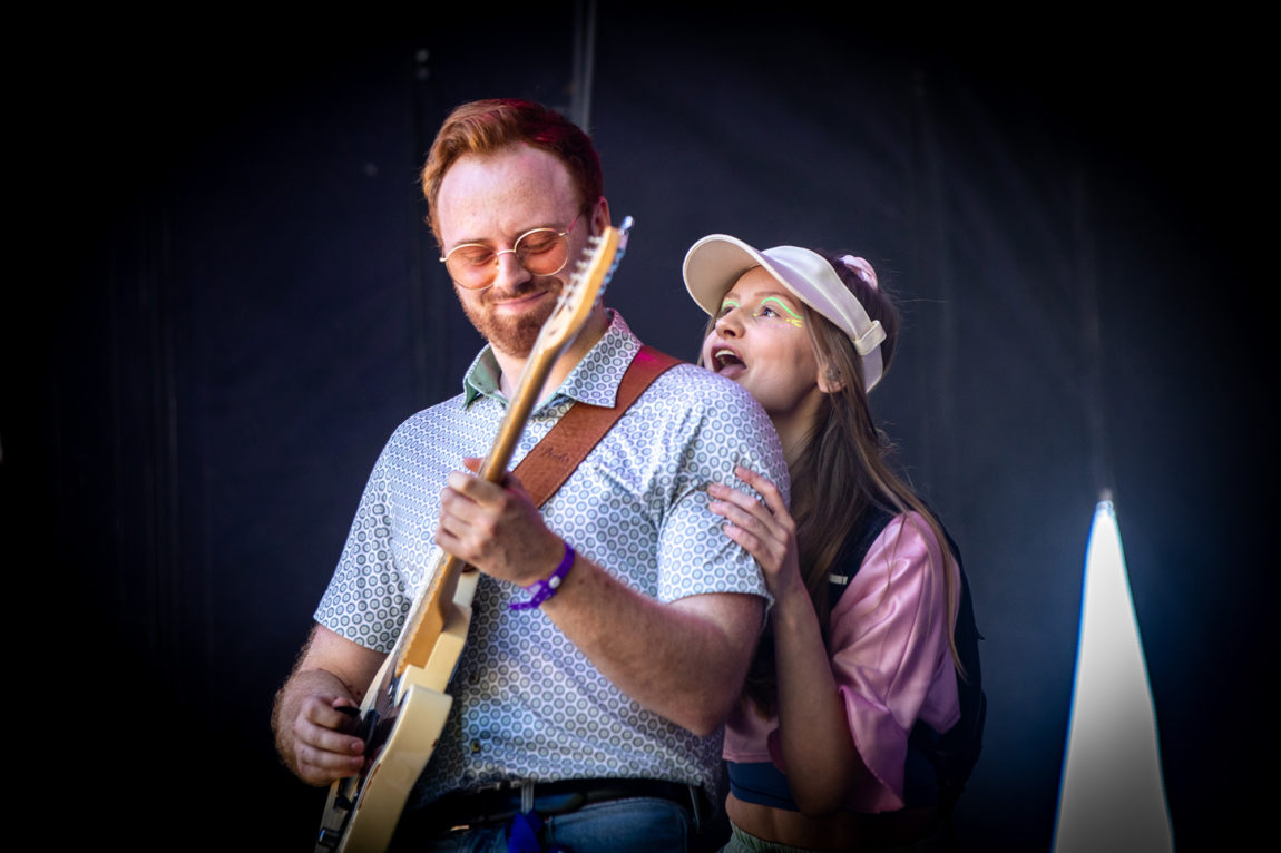 Michelle Ullestad står på scenen og holder rundt sin gitarist Martin Borge. Han har på seg en mønstrete skjorte og solbriller. Hun har på skyggelue og rosa drakt.