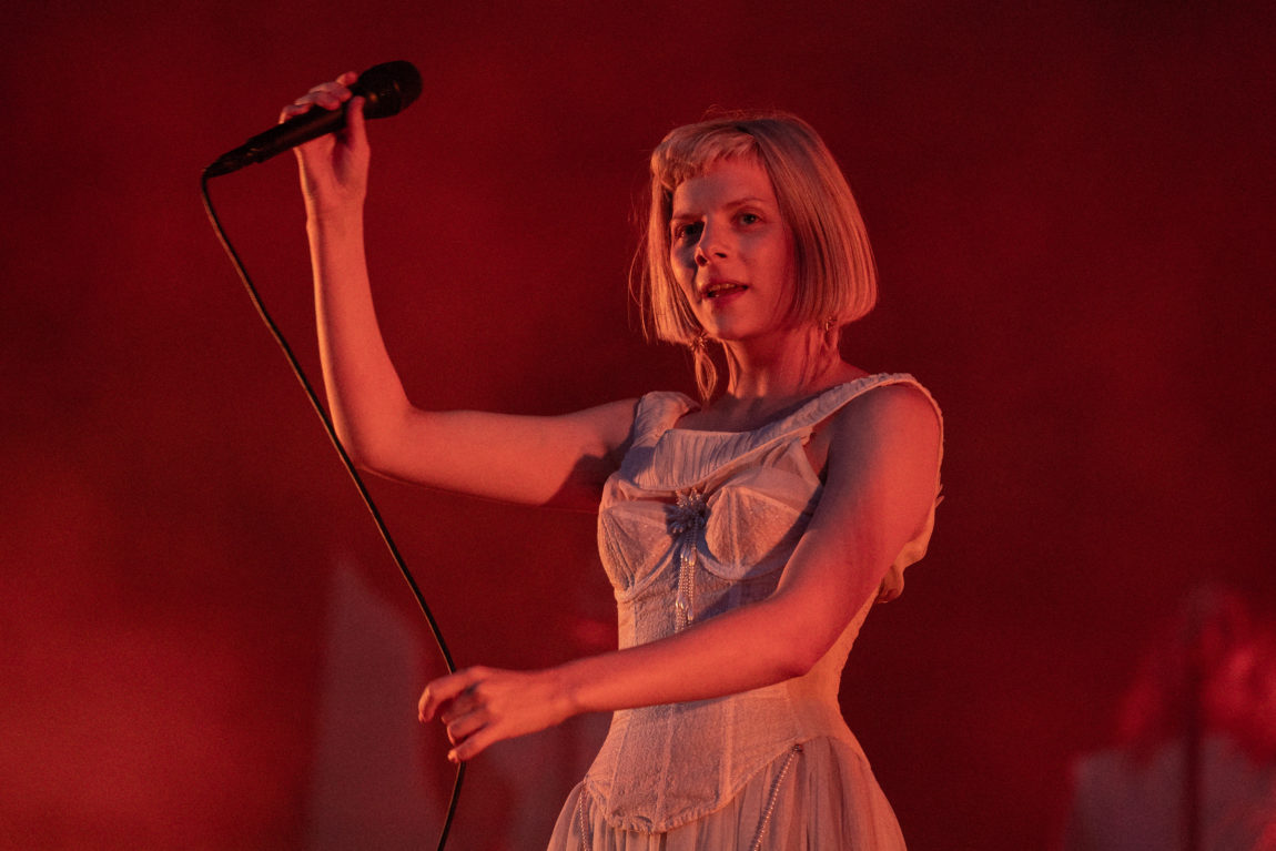 Et nærbilde av Aurora i en hvit kjole. Hun holder opp en mikrofon og i bakgrunnen er veggen rød
