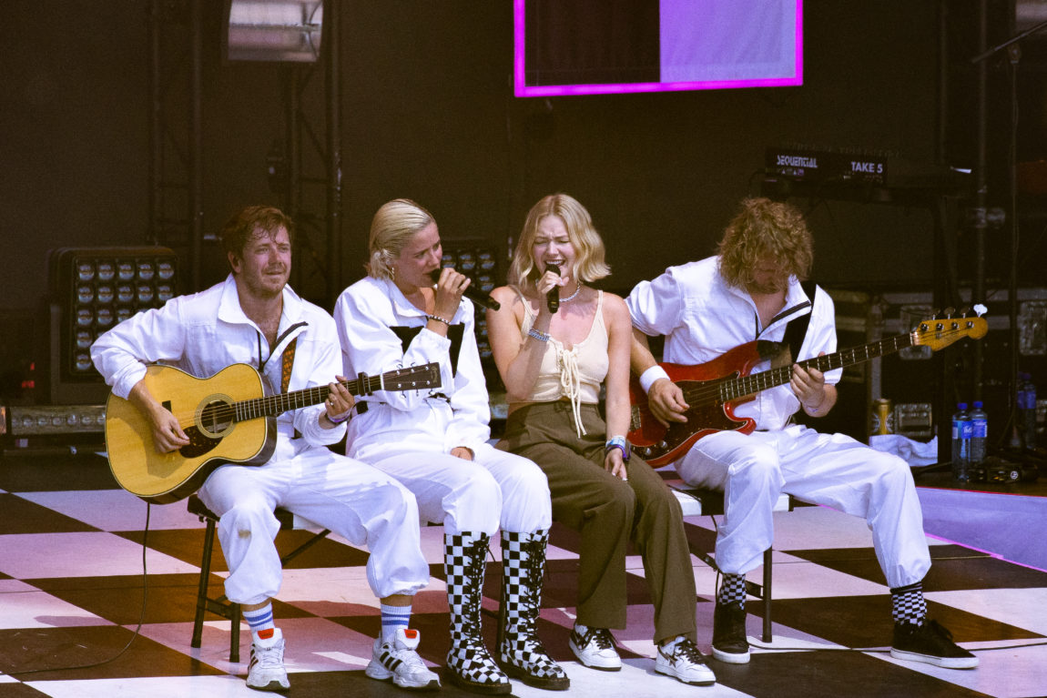 To gitarister i hvite kjeledresser sitter på hver sin side av Dagny og Astrid S.