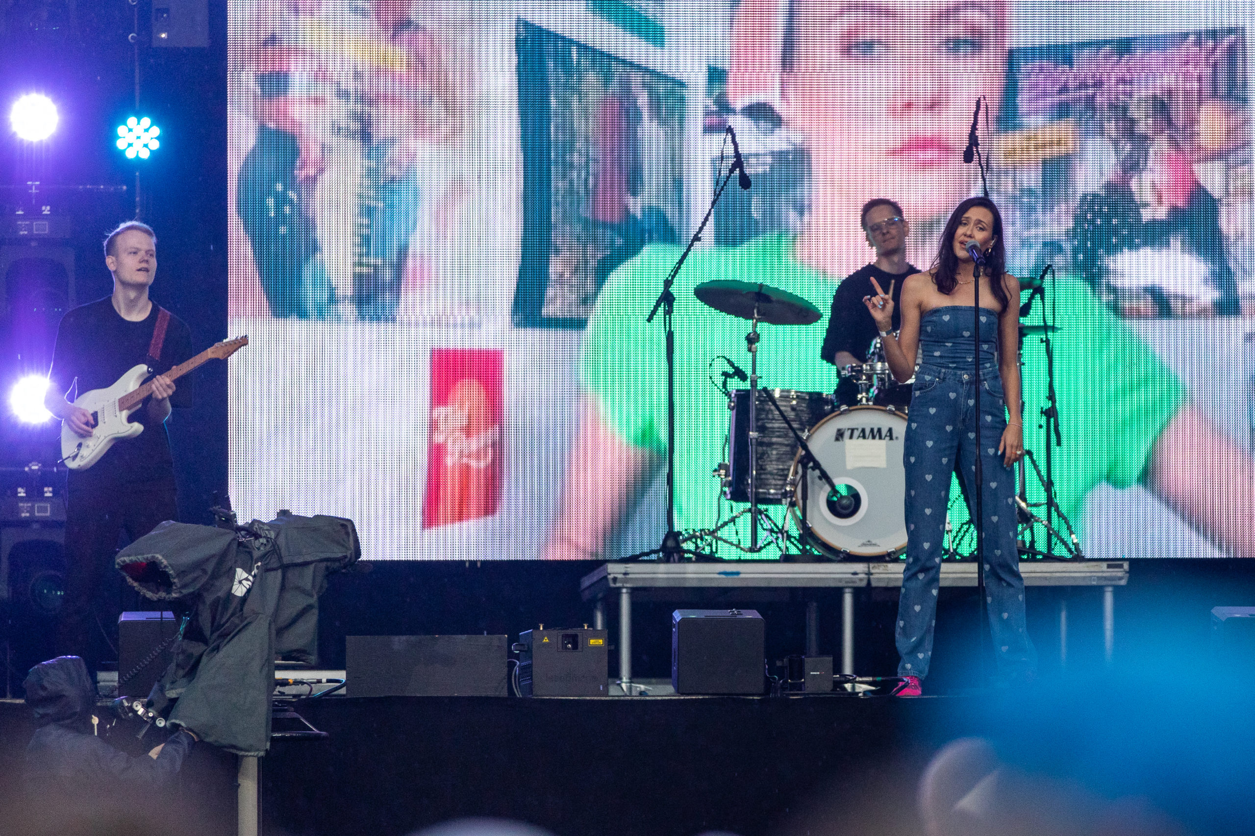 Synne Vo står på scenen og synger i sin hjerte one-piece. Hun holder en finger til værs. I bakgrunnen ser vi en gitarist og en trommis. Bak der igjen er det en skjerm hvor vi ser et bilde av Synne i en grønn t-skjorte.