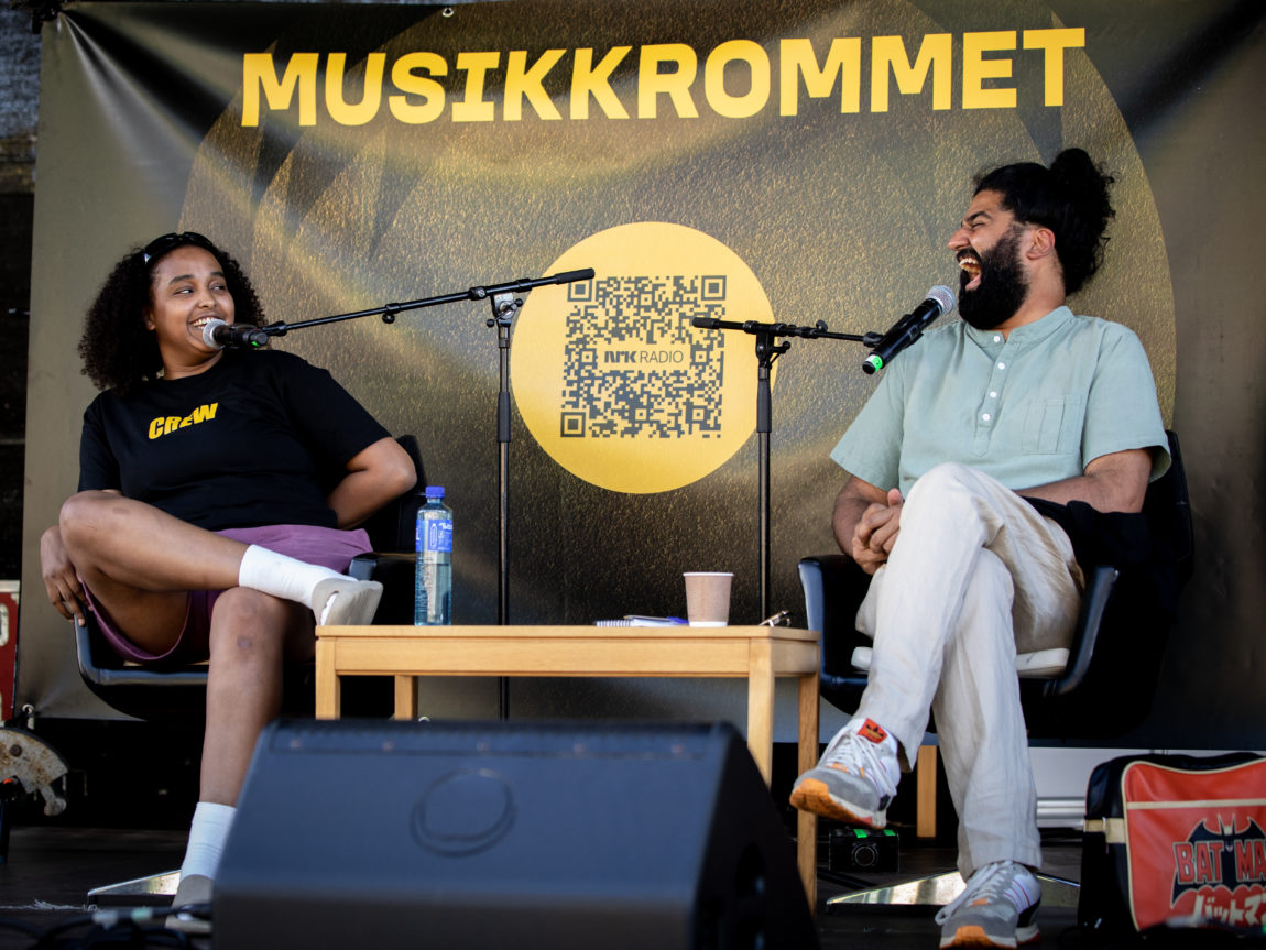 Musti sitter i en stol og prater med Sandep Singh. Musti har på seg en svart t-skjorte, har hvote sandaler og smiler i mikrofonen der hun sitter. Sandeep har på seg en beige bukse og skjorte. Han ler høyt mens han ser på Musti. 