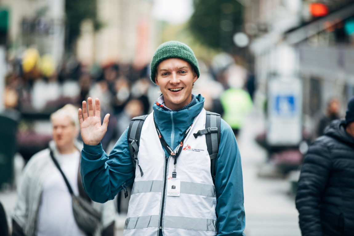 Mathias smiler til kamera mens han holder oppe en hånd og vinker. Han er lys i huden, har på seg en grønn lue som dekker til det blonde håret, en blå regnjakke, sekk og en hvit Leger uten grenser-vest på seg. Rundt halsen henger et identifikasjonskort. Bak han ser vi Karl Johans gate i Oslo og masse mennesker som går i gata. 