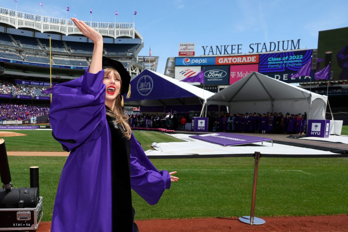 Taylor Swift har på seg en blå graduation-drakt. Hun star på en stadio med kunstgress og tribuner i bakgrunnen.