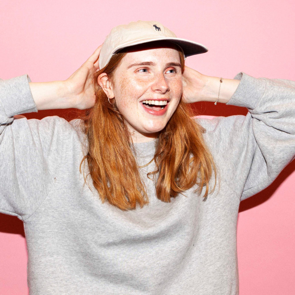 Et bilde av Anna iført grå genser og hvit caps. Hun ler foran en rosa vegg.