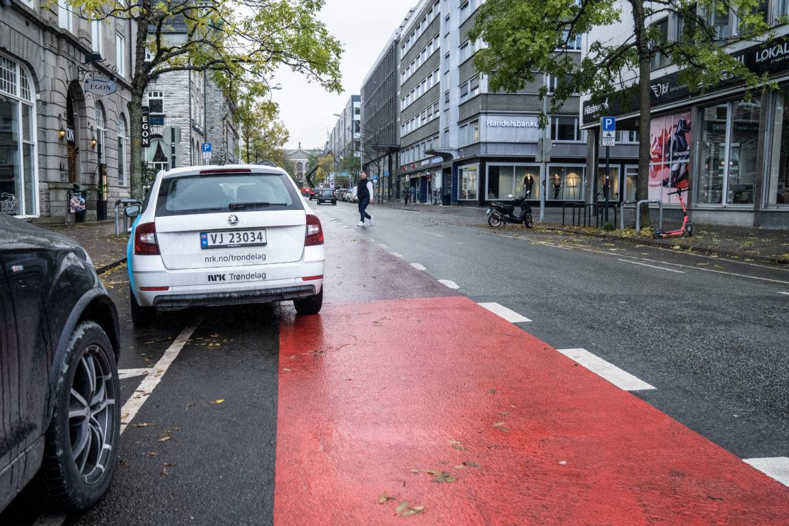 En NRK-bil står skeivt parkert utenfor en gate i Trondheim. Den står nesten parkert i et rødt sykkelfelt. Foran er en handlegate. Og en Egon-restaurant. NRK-bilen er hvit, med blå logo.