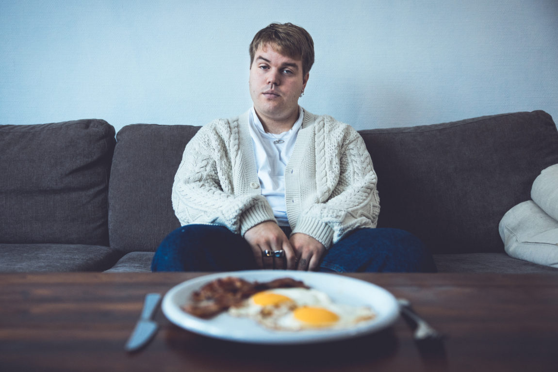 Ramón sitter i en sofa. Han ser i kamera og hviler hendene i fanget. Han har på seg hvit trøye, blå bukser og en lys cardigan. På bordet foran han er det et fat med egg og bacon.