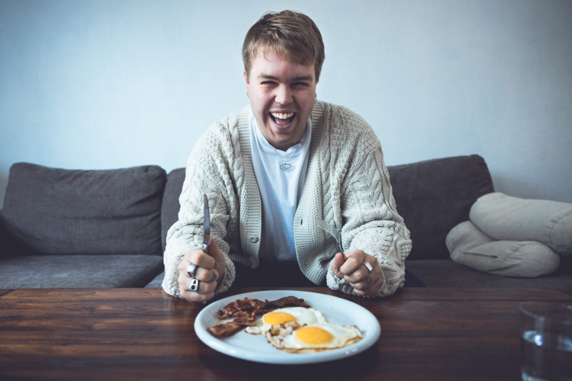 Ramón sitter i en sofa. Han har på seg hvit trøye, blå bukser og en lys cardigan. Han har bestikk i hendene og lener seg over et fat med egg og bacon mens han ler.