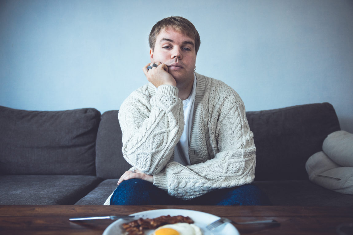 Ramón sitter i en sofa. Han ser i kamera og støtter hodet på én hånd, mens den andre hviler på låret. Han har på seg hvit trøye, blå bukser og en lys cardigan. På bordet foran han er det et fat med egg og bacon.