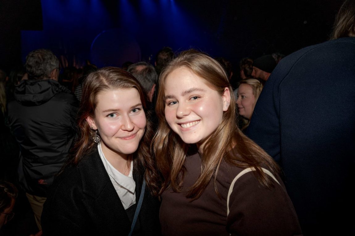 To jenter med brunt hår smiler til kamera. Hei står med menneske bak seg. Det er eit svakt belyst rom med blått lys lenger bak seg. 