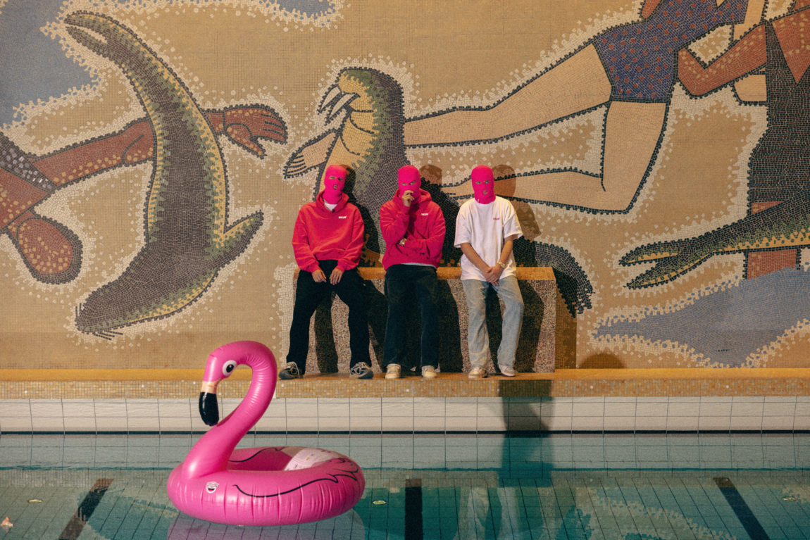 Ballinciaga sitter ved en bassengkant. Bak dem er det er digert veggmaleri og på bassenget foran dem flyter en rosa, oppblåsbar flamingo.