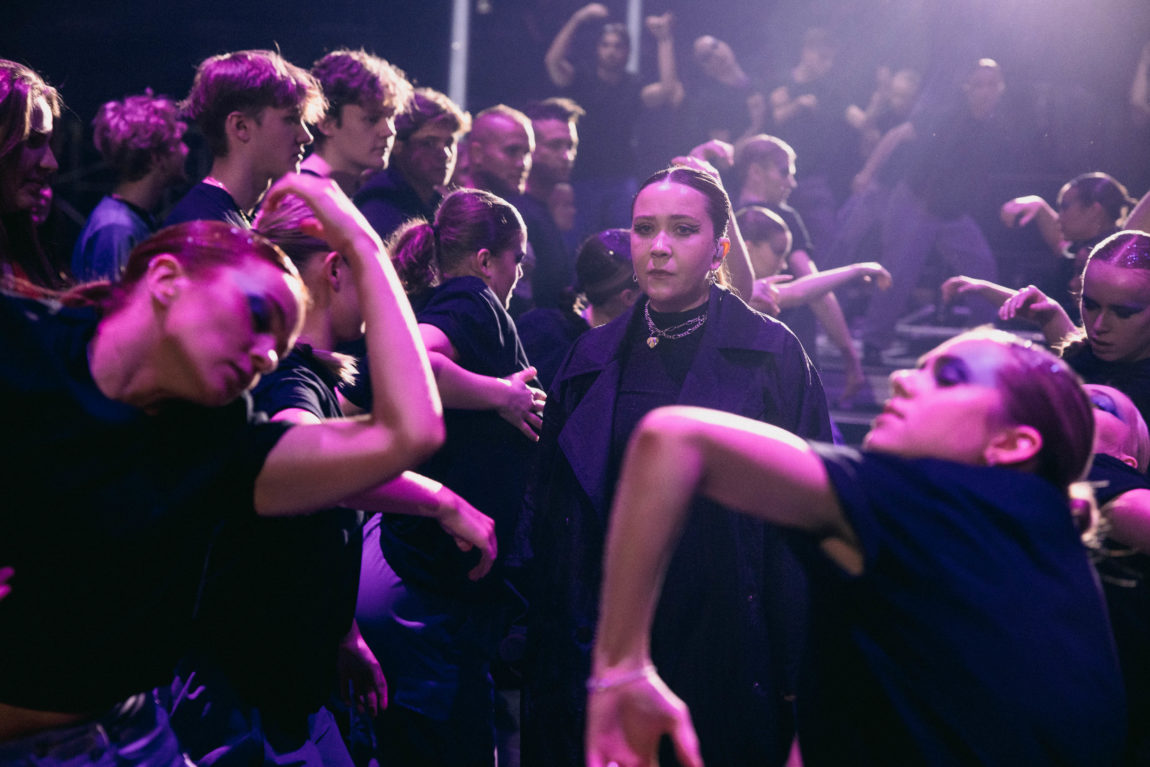 Emilie Nicola ser seriøst inn i kamera. Hun har på seg en svart dressjakke og rundt hun står det mange dansere.