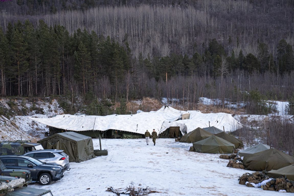 Skog og trær utan blader omsluttar  ein parkeringplass. På bakken er det snø. To personar kjem gåande over plassen. Dei er eit godt stykke unna. Bak dei, dekker ein kvit presenning ei klynge med grøne telt. Det står eit par bilar parkert til venstre i bildet. Til høgre i bildet er det slått opp små grøne telt. Utanfor telta ligg det brune sekkar i små haugar.  