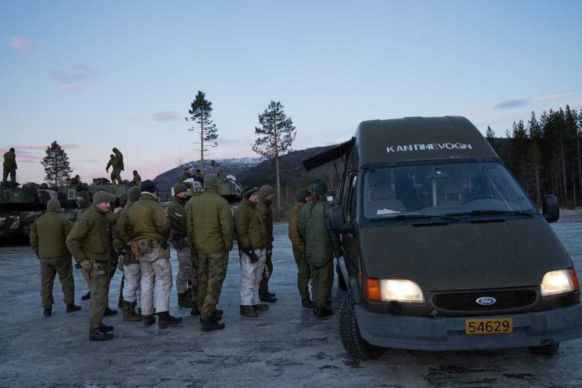 Ei grøn varevogn står oppstilt på ein parkeringsplass. Over frontruta står det skrive "kantinevogn" med kvite trykkbokstavar. Ei luke står open på den eine langsida til bilen. Foran luka har det danna seg ei kø av soldatar. På bakken er det is. I bakgrunnen står stidsvogner stilt opp og det er soldatar som går på taket. Litt skog og fjell i horisonten. Himmelen er blå med litt rosa skyer. 