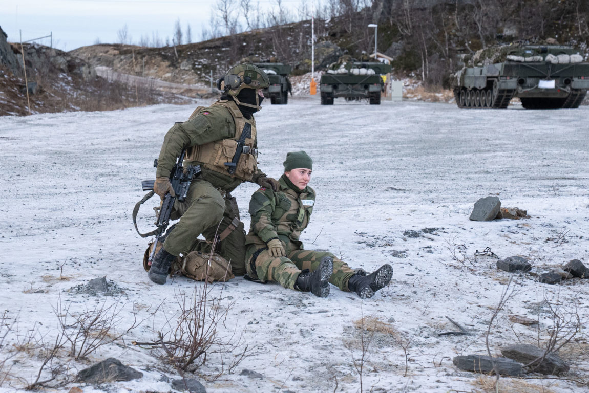 Ein soldat med eit maskingevær i den eine handa har lagt den andre handa på skuldra til ein soldat som sit på bakken. Soldaten på bakken blir holdt igjen. Rundt låret er det festa ein svart stropp og soldaten held seg til låret. Blikket til soldaten er tomt og ho stirrar rett ut i lufta. Soldaten som held igjen knelar på bakken, men verkar vere på veg ein stad. Det er som som han ser ut av bildet der han er opptatt med noko anna. I bakgrunnen står tre stridsvogner stilt opp. 