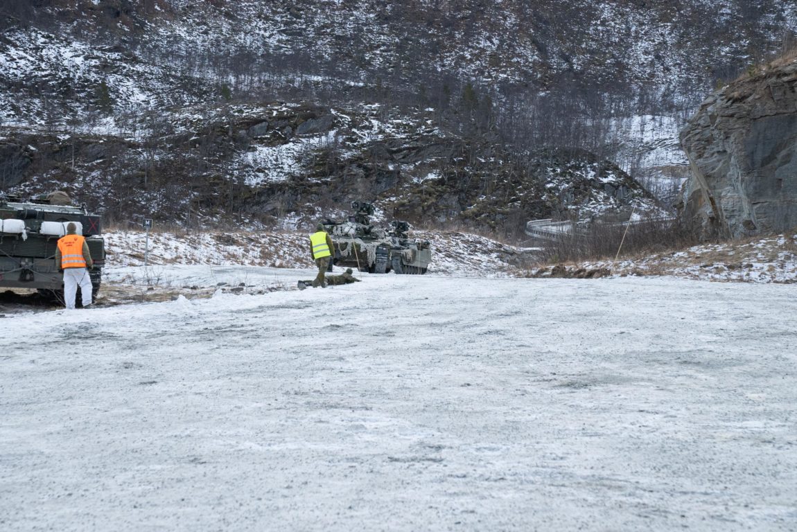 Oversiktsbilde av ein situasjon. Ein person i grøne militørklede og gul refleksvest står ved foten av ein soldat som ligg på bakken ut mot vegen. To stridsvogner kjem køyrande opp vegen. Til venstre i bildet står det ein person i grøn militærjakke og kvit bukse og oransje refleksvest ved bakenden av ei stridsvogn. 