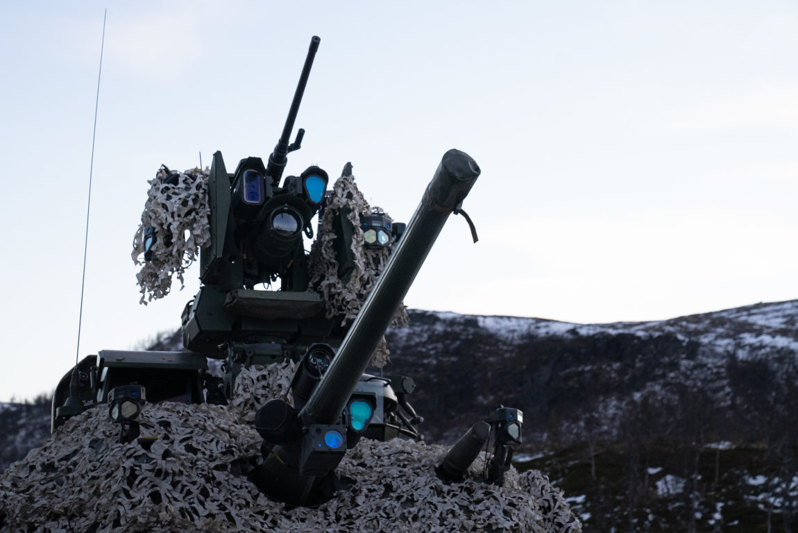 Nærbilde av ein kanon på taket av ei stridsvogn. Rundt kanonen er det festa svarte boksar, linser og prismer. Boksane har blått glass. Dette er sensoarar og laser. Ein grå presenning ligg rundt kanonen. Teksturen er ruglete og kan minne om mose. I bakgrunnen er fjellet dekt av eit lett lag snø. Himmelen er lys. 