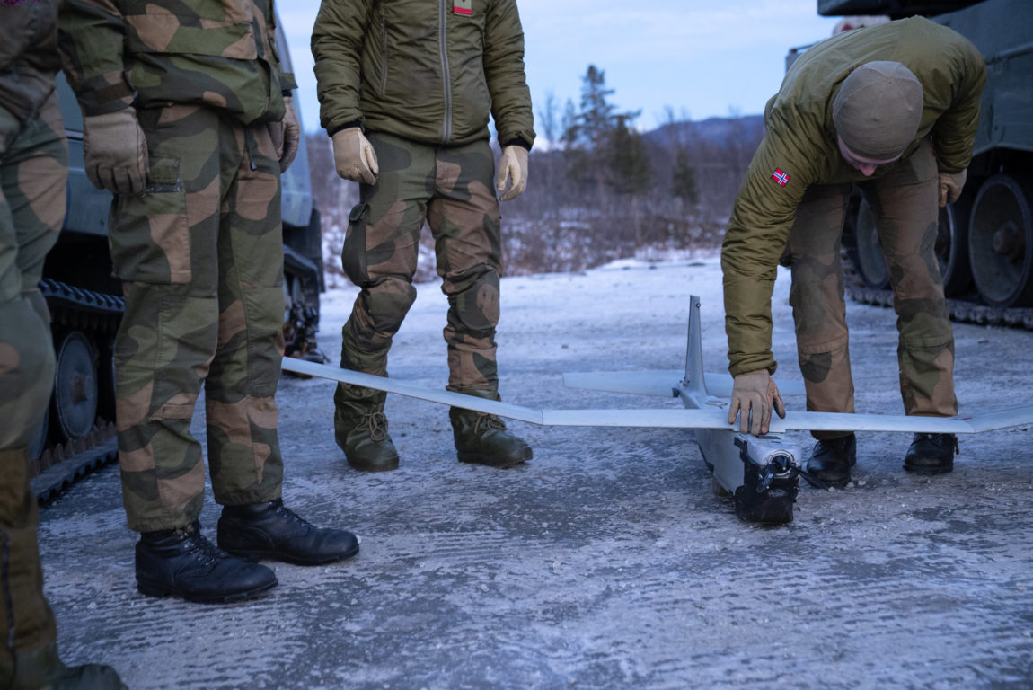 Eit grått lite fly står på bakken. Ein soldat bøyger seg over flyet og støttar det med handa. Runst flyet står tre andre soldatar, men vi ser berre beina. Det grå flyet er ei drone. 