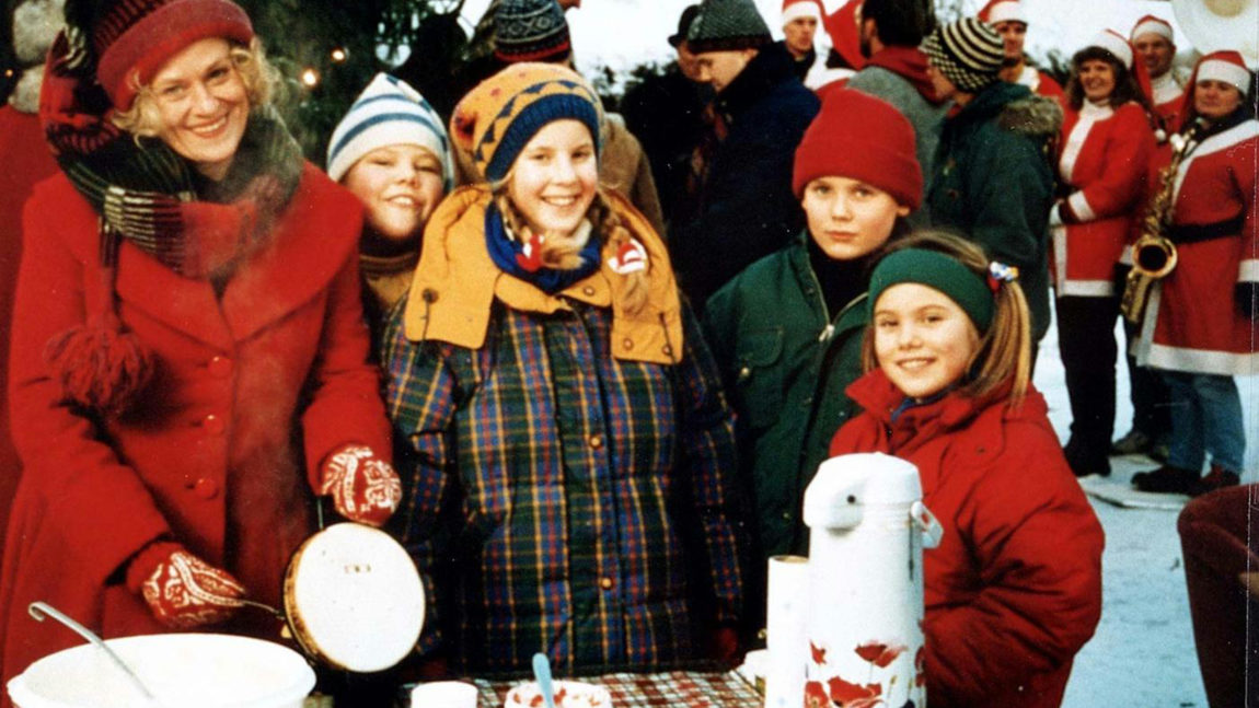 Et bilde fra julekalenderen Amalies Jul. Flere står rundt et bord med røde julejakker og nisseluer.