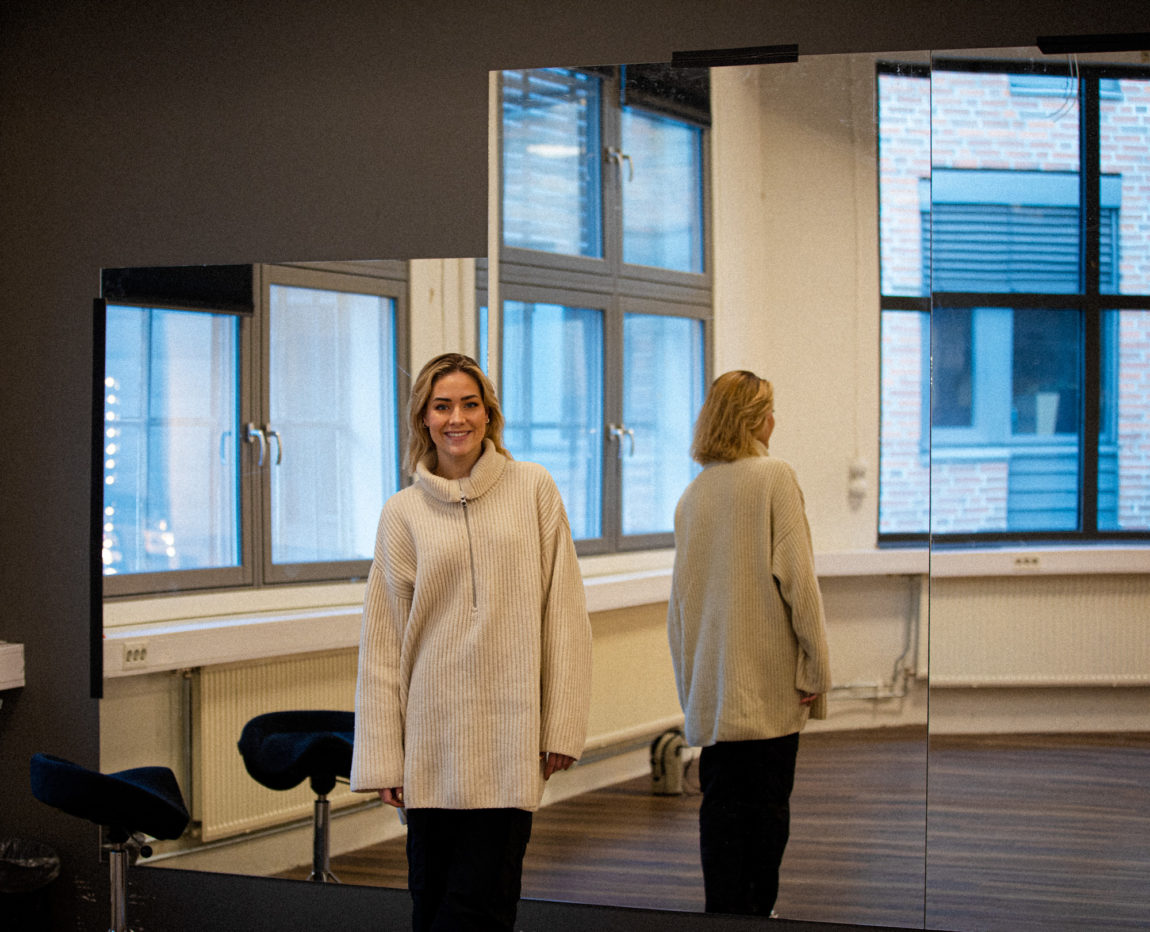 Helene smiler litt på avstand til kamera. Hun står foran store speil inne i den ene salen i dansestudioet sitt. Hun har blondt, løst hår. Hun har på seg en stor, hvit ulljakke, med sølvarget glidelås og høy hals. Hun har på svarte bukser. Ryggen hennes ser man i speilene hun står foran, og man skimter også store vinduer. 