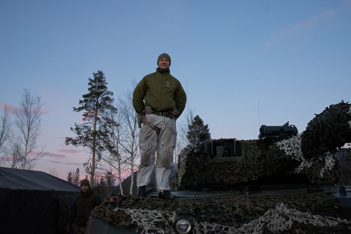 Ein ung mann står oppå eit panser. Han har armane på ryggen og smiler ned mot kamera. Han har på seg ei grøn hue og jakke. Buksene er kvite, men skitne. Rundet det eine låret er det festa ein brun stropp som går til eit våpenhylster. Panseret han står på er dekt av eit grønt, ruglete nett med kvite detaljar. Eit kanonløp går ut frå eit lite rundt hus på toppen avpanseret. Himmelen er blå, men dei rosa skyene vitnar om solnedgang. 