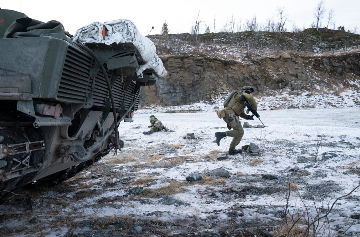 Delar av bakenden på ei stridsvogn er synleg i forgrunnen. Ein soldat med gevær er i full firsprang vekk frå vogna. I bakgrunnen ligg ein soldat på bakken som drar seg mot vegen. 