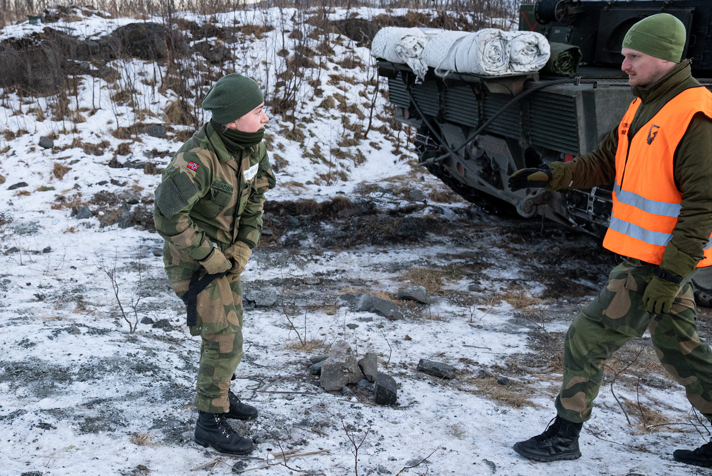 Ein mann i grøne militærklede og ein stor sjølvlysande organsje vest med refleks kjem inn frå sida. Den eine armen er heva som om han skal seie noko. Blikket er konsentrert. Han går mot ein soldat som løsnar ein svart stropp rundt låret, medan ho ser mot mannen i den oransje vesten. I bakgrunnen stikk ein bakende av ei stridsvogn fram. Bakken er steinete og dekt av eit tynt lag snø og is. 