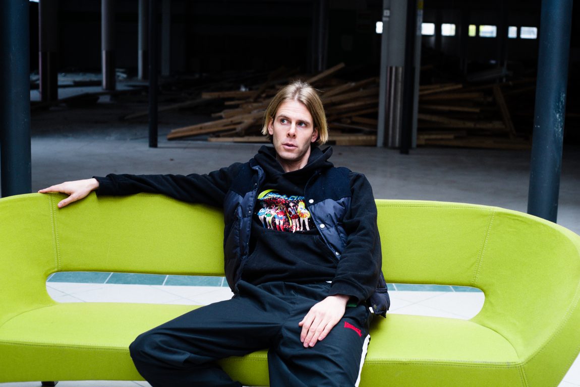 Lars Vaular har på seg en svart bukse, hvite joggesko og en svart hettegenser. Han sitter i en grønn designersofa.