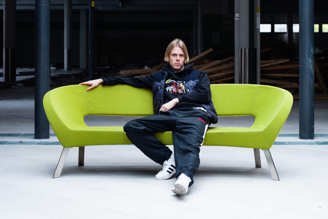 Lars Vaular sit i ein grøn sofa og ser i kamera. Han er kledd avslappa, og lokalet han er i er under renovering.