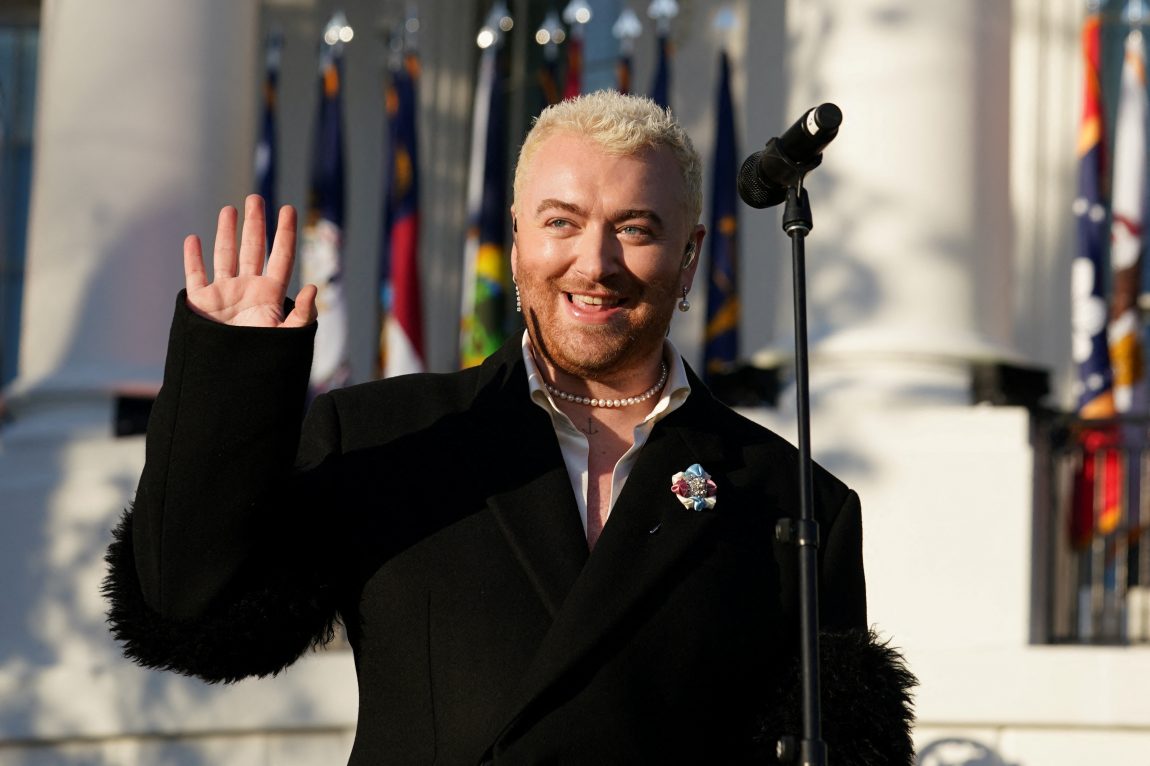 Sam Smith har hvitt hår og en svart dresskåpe. Hen smiler og vinker til publikum.