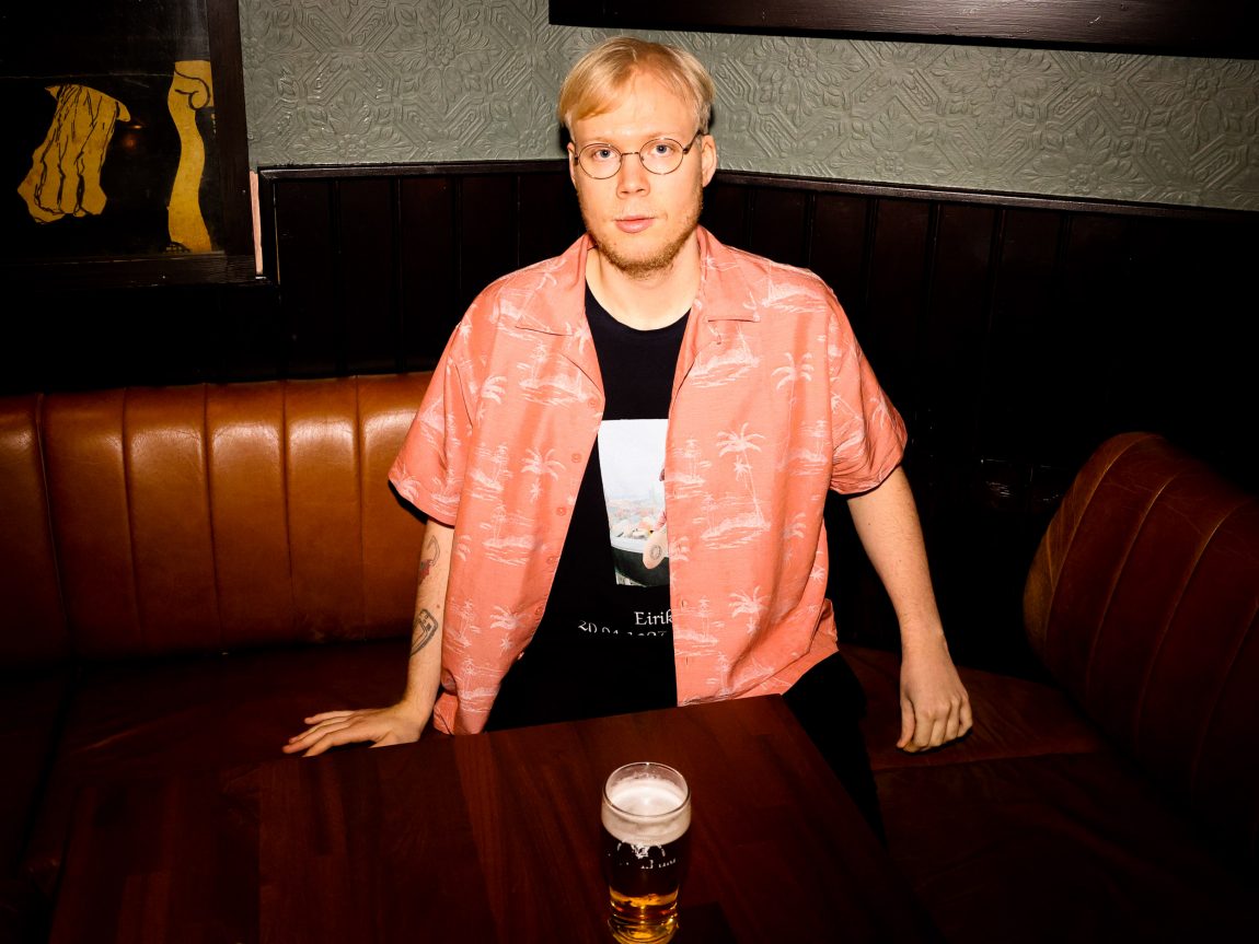 Eirik sit i eit hjørna på den brune puben. Han ser alvorleg i kamera og har hendene ned langs sida i skinnbenken. Framfor han står eit glas med øl. 