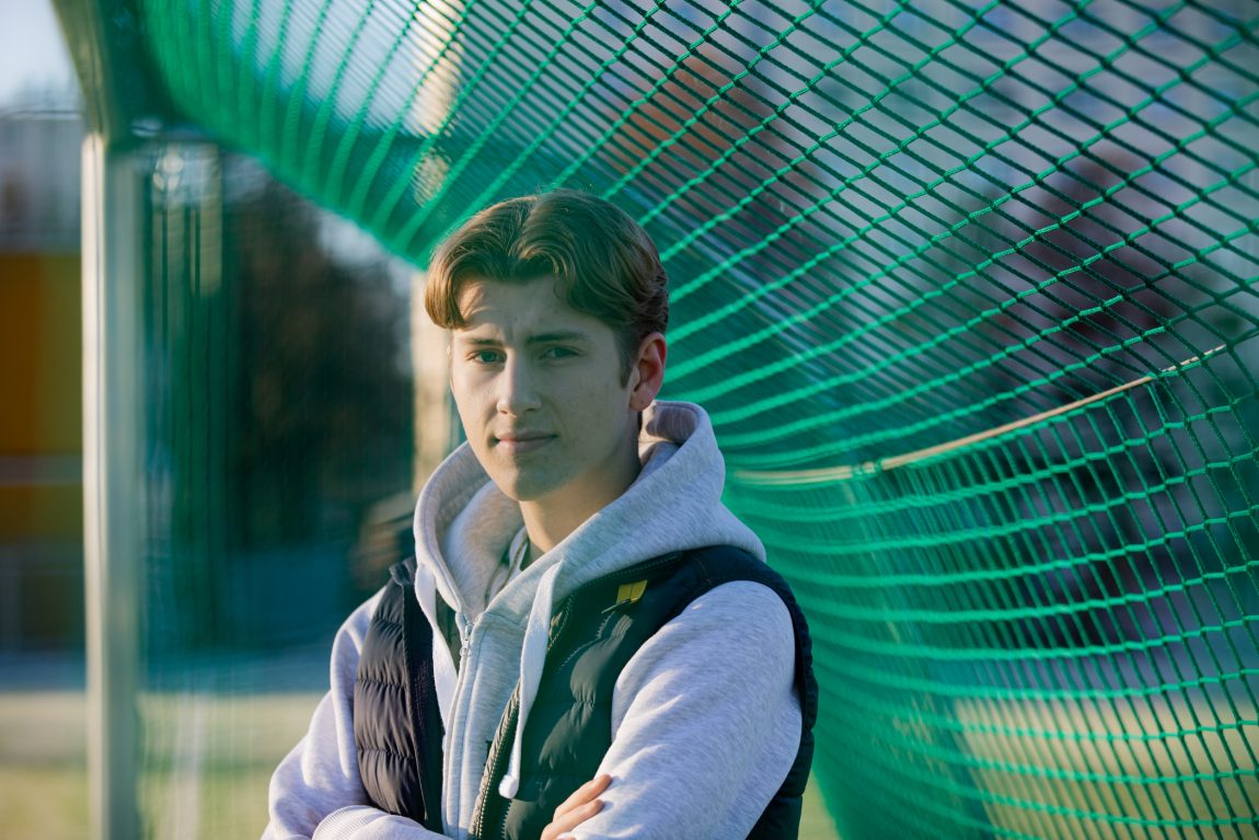 Isaac kikker alvorlig inn i kamera, med armene i kors over den mørkeblå boblevesten og lysegrå hettegenseren. Han står inne i et fotballmål, og der \\t er grøn rundt ham. 