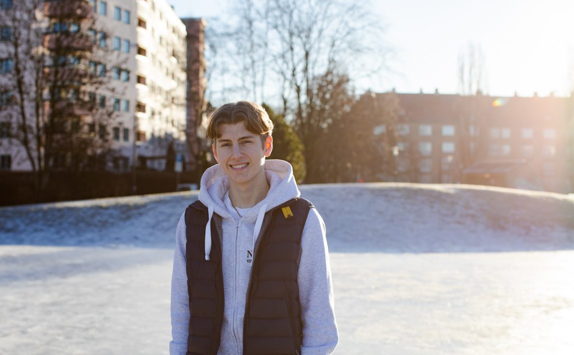 Isaac smiler i kamera. Det er solfyllt rundt han, og solen skinner ekstra fra høyre siden. Han har på seg en mørkeblå boblevest over en lysegrå hettegenser. Han har blond midtskill. Bak han er en snødekt hau, og en solinnrammet bligblokk. 