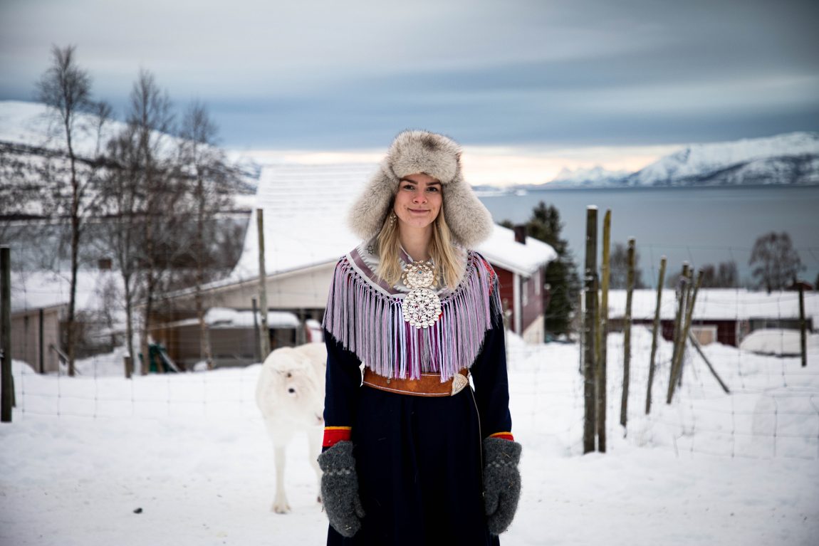 Kine kikker inn i kamera og msiler forsiktig. Hun har på seg koften sin, og en stor pelslue med flapper. Bak henne ser man deler av huset, og store hvitkledde fjell. Sola bryter såvidt med en skydekt, blå himmel i horisonten mellom fjellene og havet. Rett bak Kine ser man en hvit, liten rein