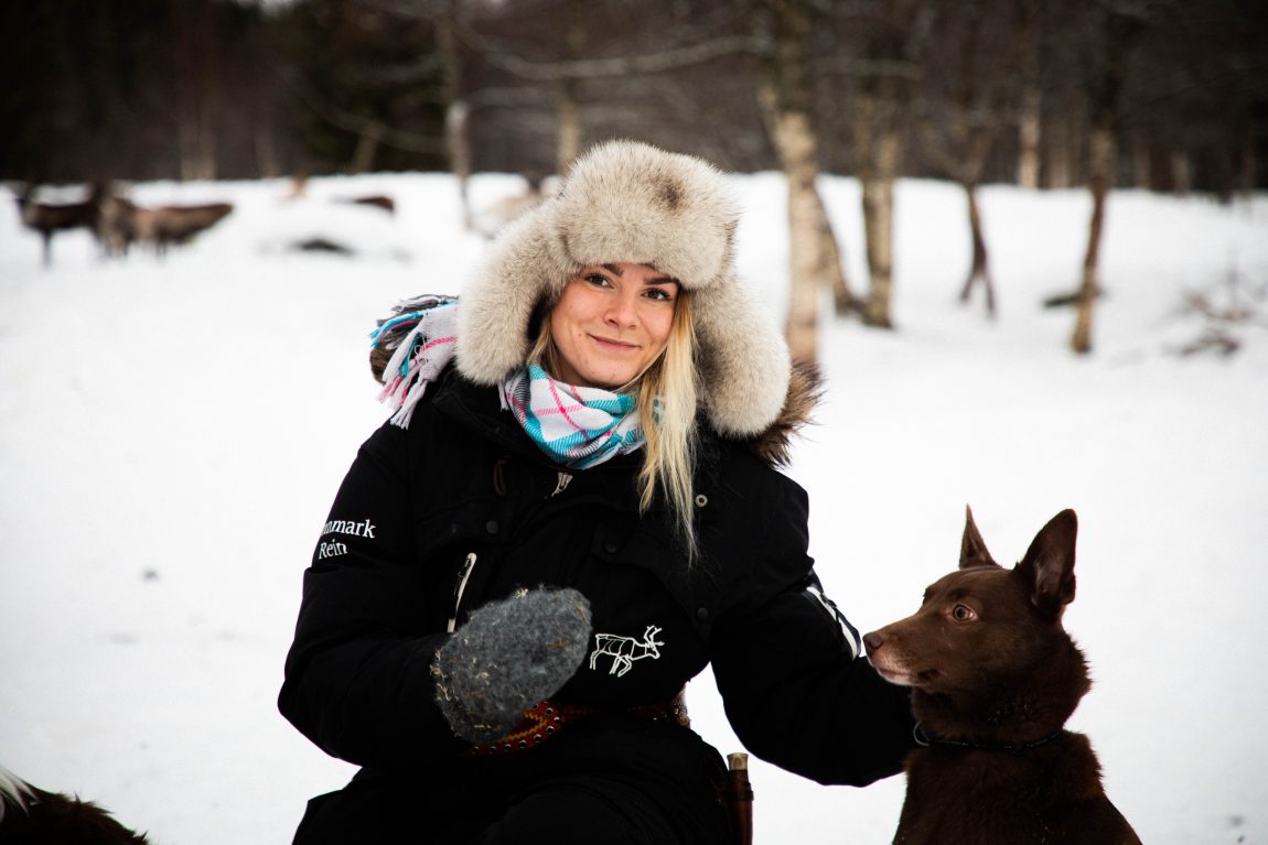 Kine kikker smilende i kamera. På hodet har hun en stor pelslue med flapper. Hun har på seg et blått, hvitt - o g rosarutete skjerf. Ved siden av henne sitter hunden Ruste som skikker spørrende på hånden henens, som har en grå vått på. Litt av Kine sitt blonde hår stikker ut av lua. 