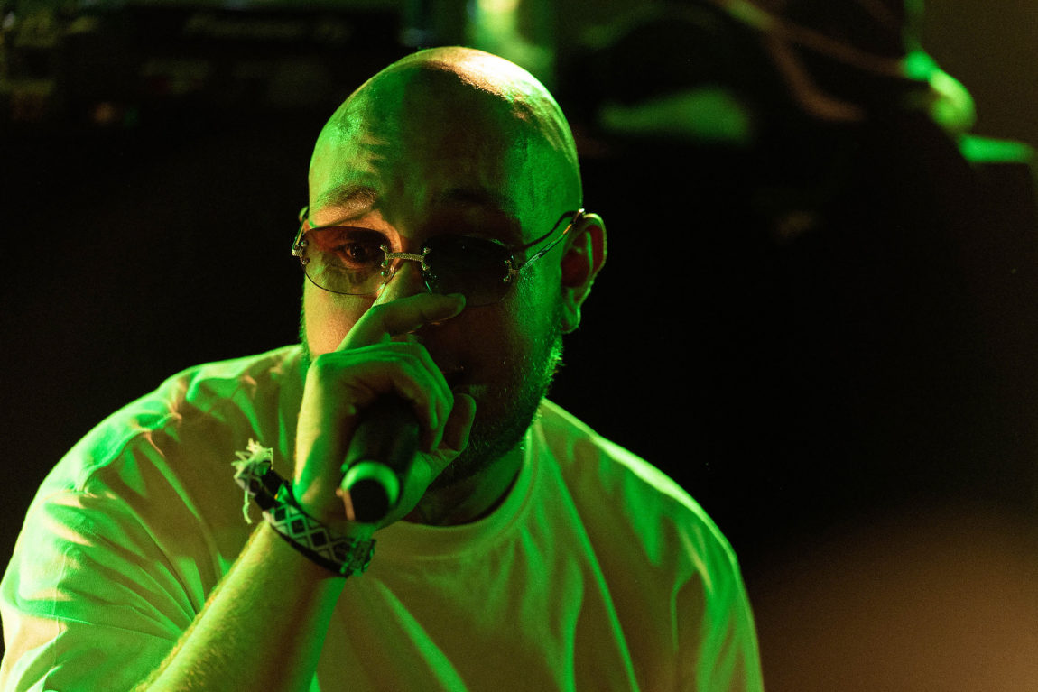 Tyr har på seg briller og en hvit t-skjorte og rapper inn i en mikrofon.