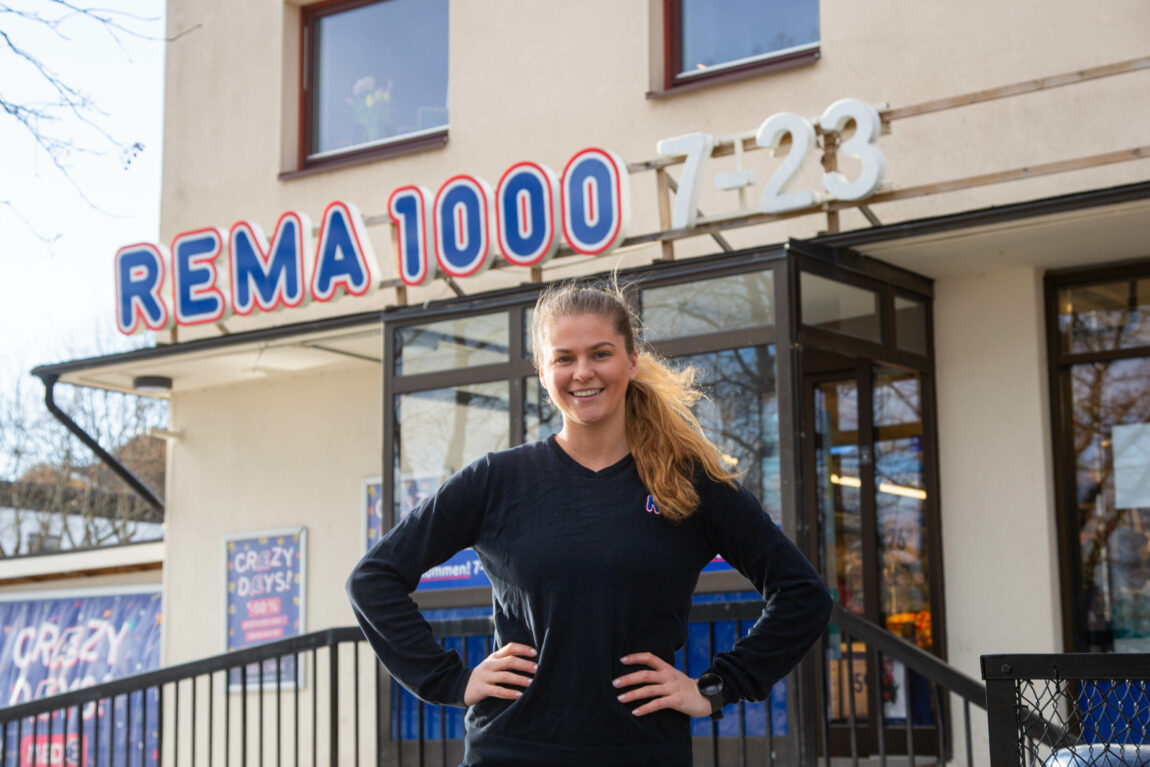 Cathrine står med hendene på hoftene foran Rema 1000 butikken. Hun har på seg Rema 1000 uniform og har håret opp i en hestehale. 