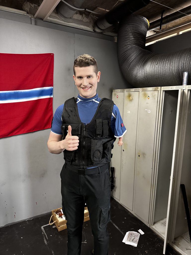 Henrik Farley er avbildet, og er iført politiaktig kostyme på.  Han smiler, venstre arm viser tommel opp, høyre arm er erstattet av en liten falsk babyarm. I bakgrunnen henger et halvt Norgesflagg, det står en skaprekke som typisk hører hjemme i en gymgarderobe, og et stort ventilasjonsrør. 