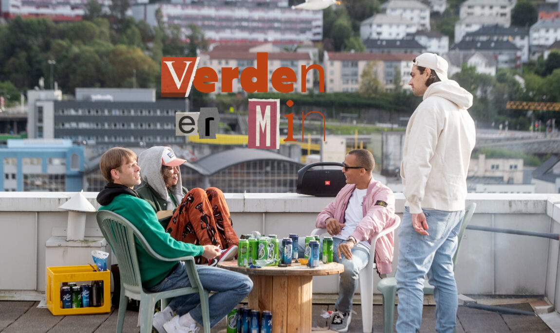 Lars Vaular, Odilie, Kamelen og han siste sitter rundt et bor med masse øl på en veranda i Bergen. Vi ser skriften «Verden er min» på skjermen. Bak er det masse boliger.