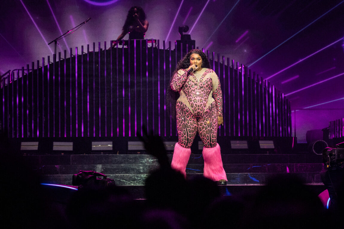 Lizzo har på seg en kjole som er rosa og grønn. Hun står på scenen i sie rosa buffalo sko.