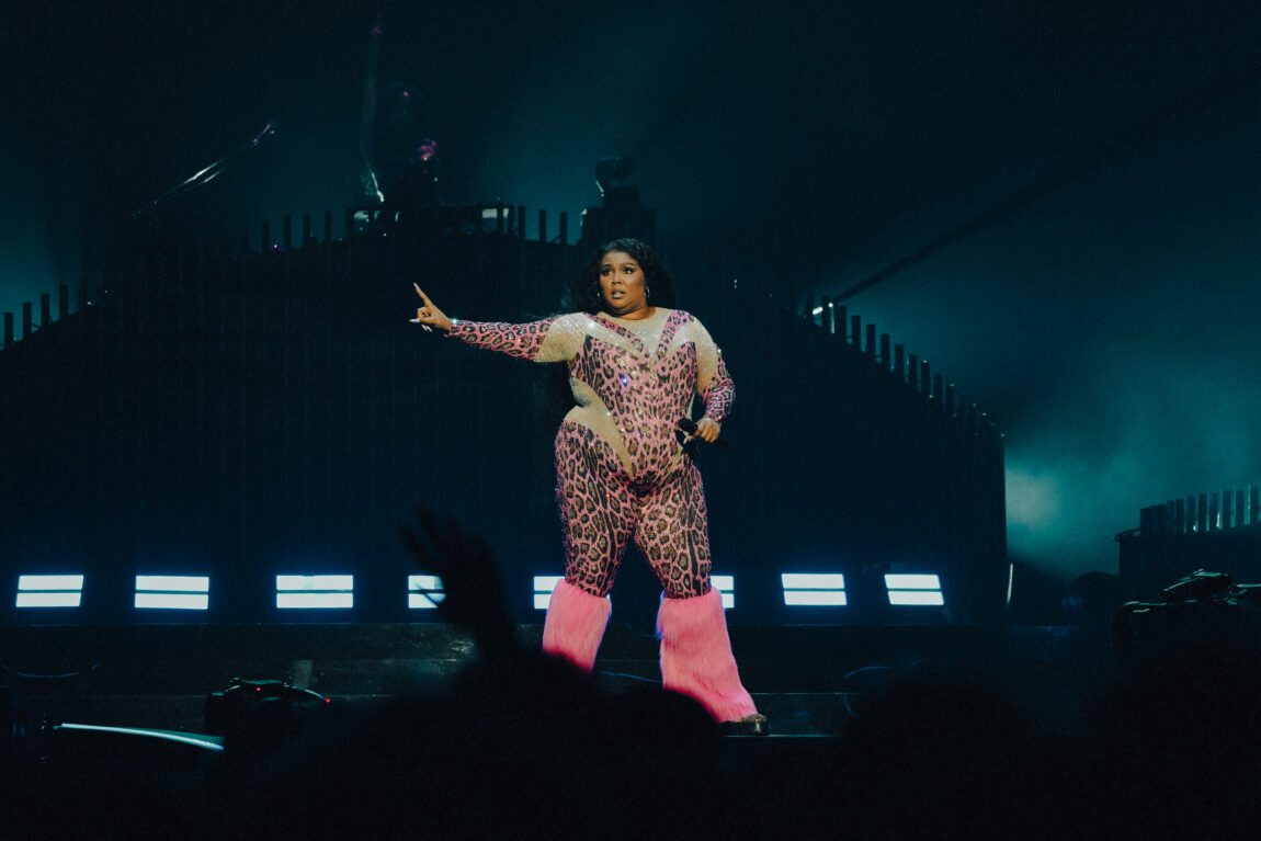 Lizzo har på seg en kjole som er rosa og grønn. Hun står på scenen og peker mot publikum.