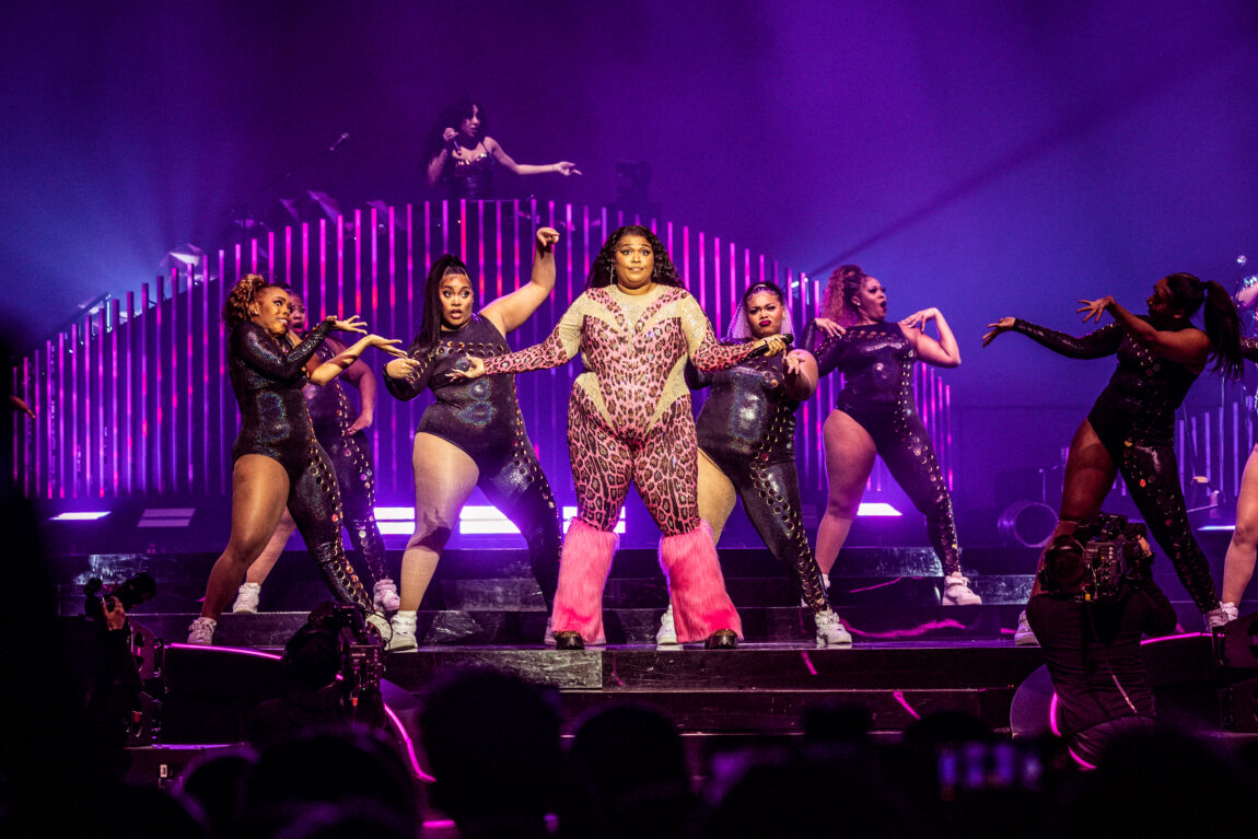 Lizzo står på scenen i en roda leopard kjole med grønne detaljer. Rundt hun står det flere dansere i lateks kjoler.