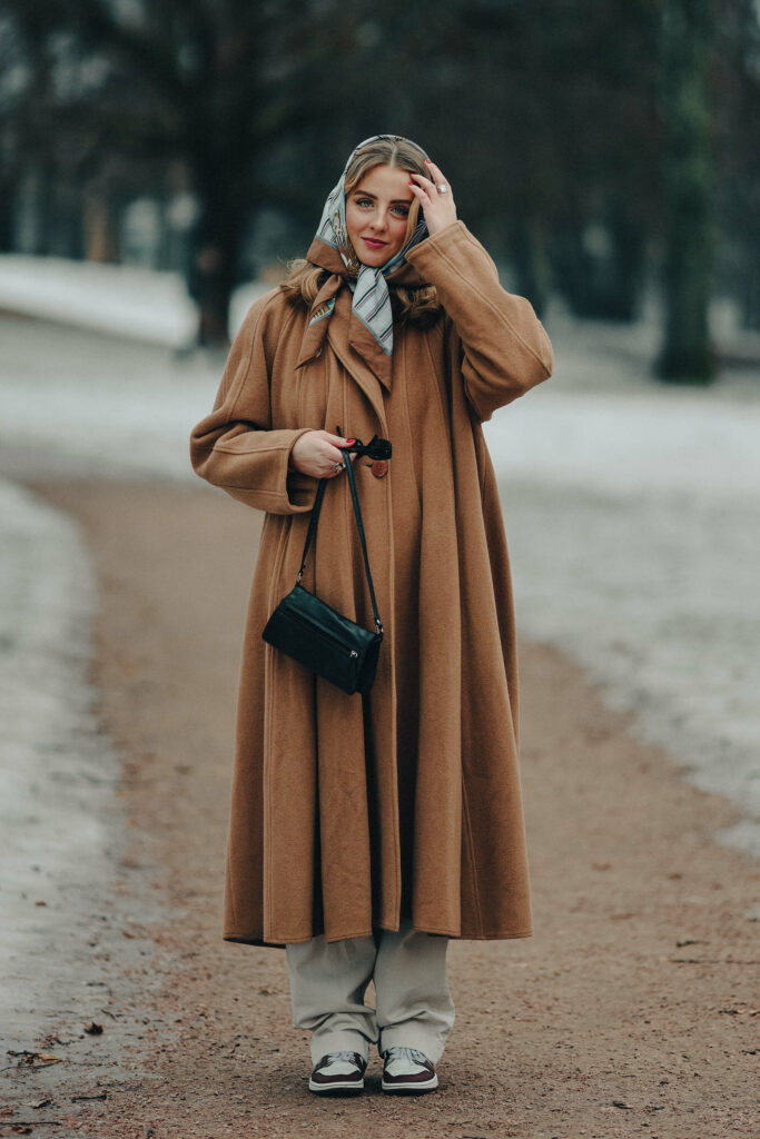 Kristin står i parken. Hun holder en veske i den ene hånden, og med den andre hånden tar hun håret bort fra fjeset. Antrekket hennes er i fokus. På hodet har hun et sjal. Kåpa er fotsid og har store knapper, buksene folder seg over skoen, hun har sneakers på, veska er svart og neglene er lakket rosa. I bakgrunnen skimter man trær og snø.