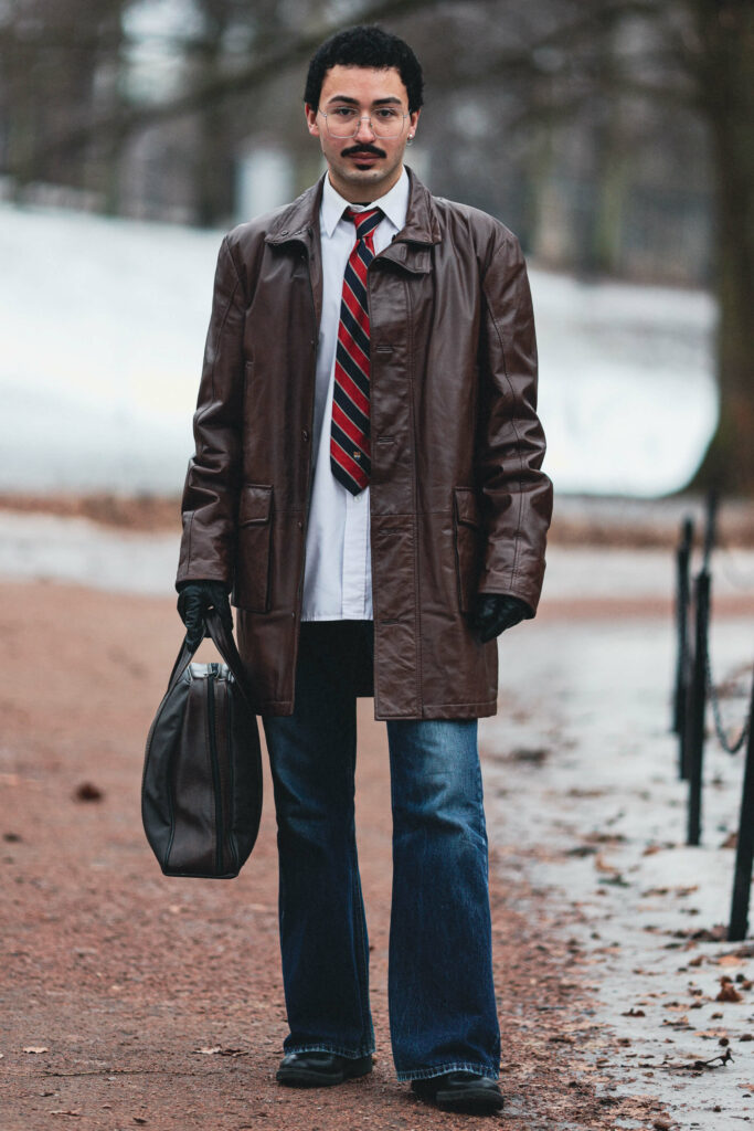 Yunus er fotografert i helfigur i parken. Han har på seg en brun skinnjakke, svarte skinnhansker, hvit skjorte, stripete rødt og blått slips, jeans og svarte sko. I hånda holder han en skinnveske. Håret hans er svart, han har briller, distinkt bart og litt skjegg, rett under underleppa. Han ser rett inn i kamera.