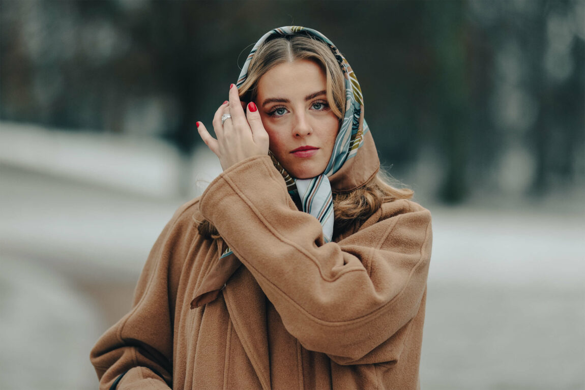 Kristin står i parken, og ser rett inn i kamera. Hun tar håret bort fra fjeset med den ene hånden. Ansiktsuttrykket er nøytralt. Knyttet rundt hodet og under haken har hun et silkesjal. 