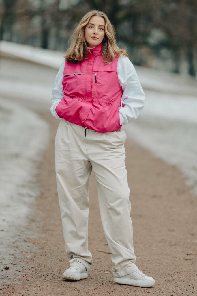 Kristin står i parken, vi ser hele henne. Hun har på seg en genser med en boblevest over i sterk rosa. Baggy bukser og hvite sneakers. I bakgrunnen skimtes en sti, snø og noen trær.