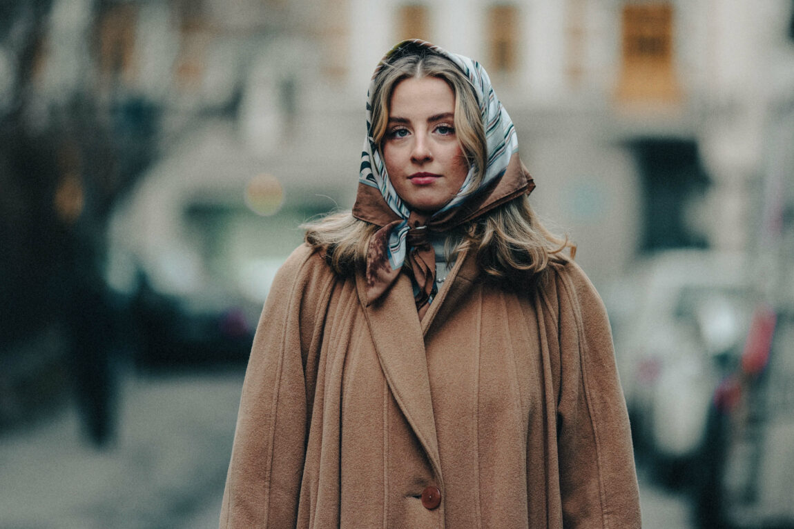 Kristin er fotografert fra livet og opp, utendørs. Hun ser rett inn i kamera, med antydning til et smil. Kristin har brunt hår som rekker henne litt nedenfor skuldrene, og rundt hodet har hun knyttet et silkesjal med striper. Kåpa er lysebryn og har store knapper. I bakgrunnen skimter man et tre og noen biler, men Kristin er i fokus. Det er tydelig at hun står i en gate.