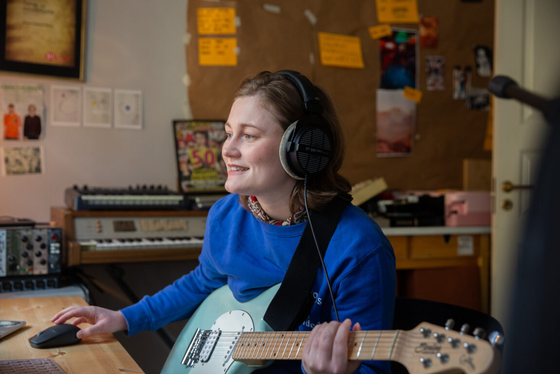 Smilende produserer Haley musikk fra hjemmestudioet sitt. Hun har på seg en blå genser og sitter med sin pastellblå gitar i fanget. 