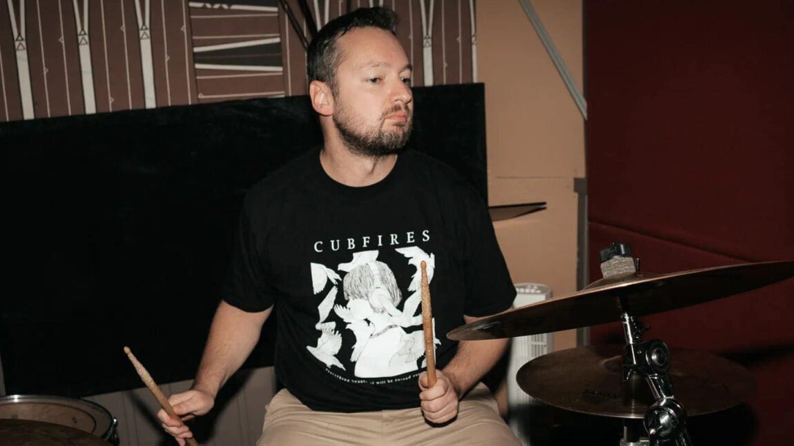 Morten Samdal sitter i svart t-skjorte og brune bukser og spiller trommer.